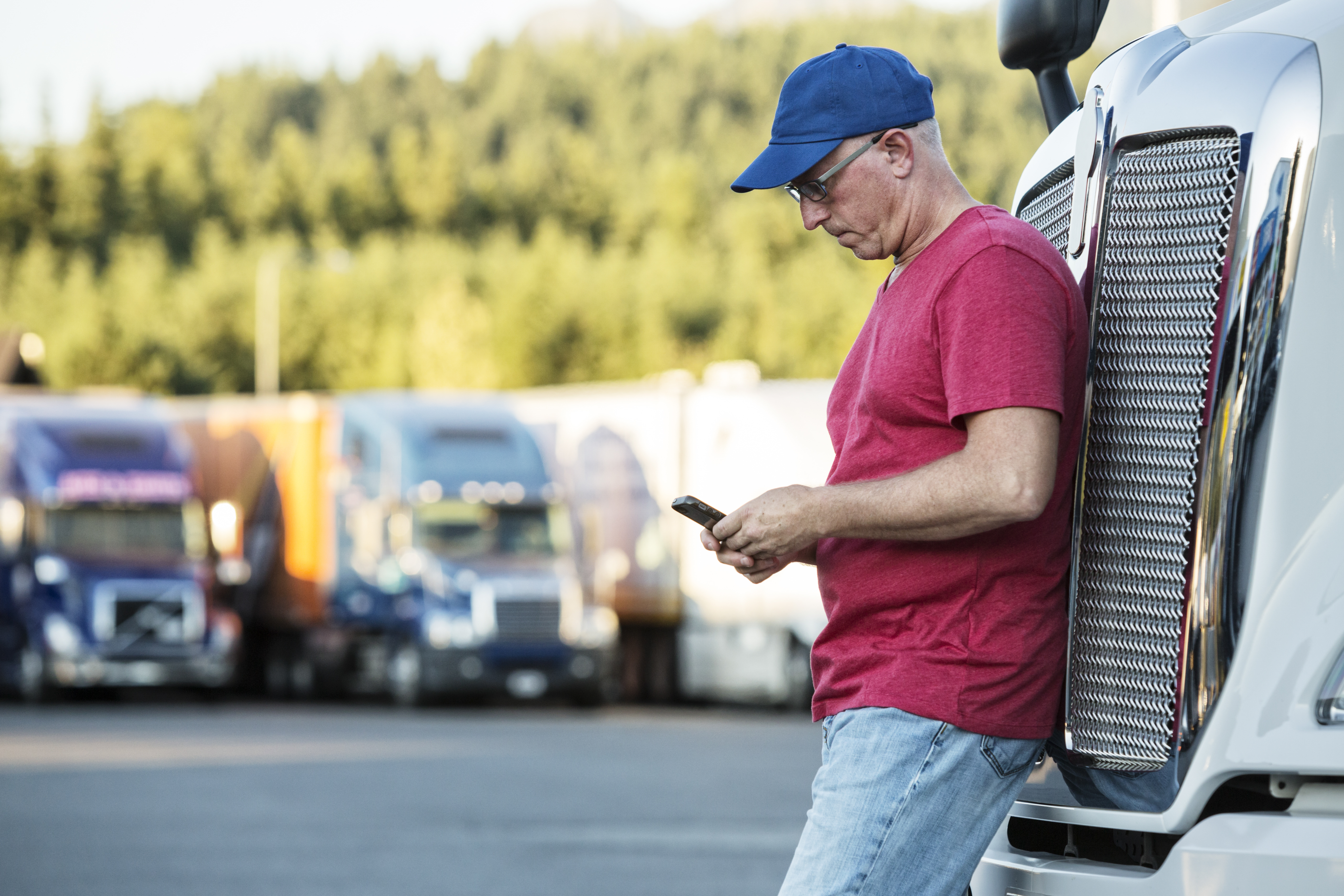 truck driver leaning up against semi truck looking down at phone in hand