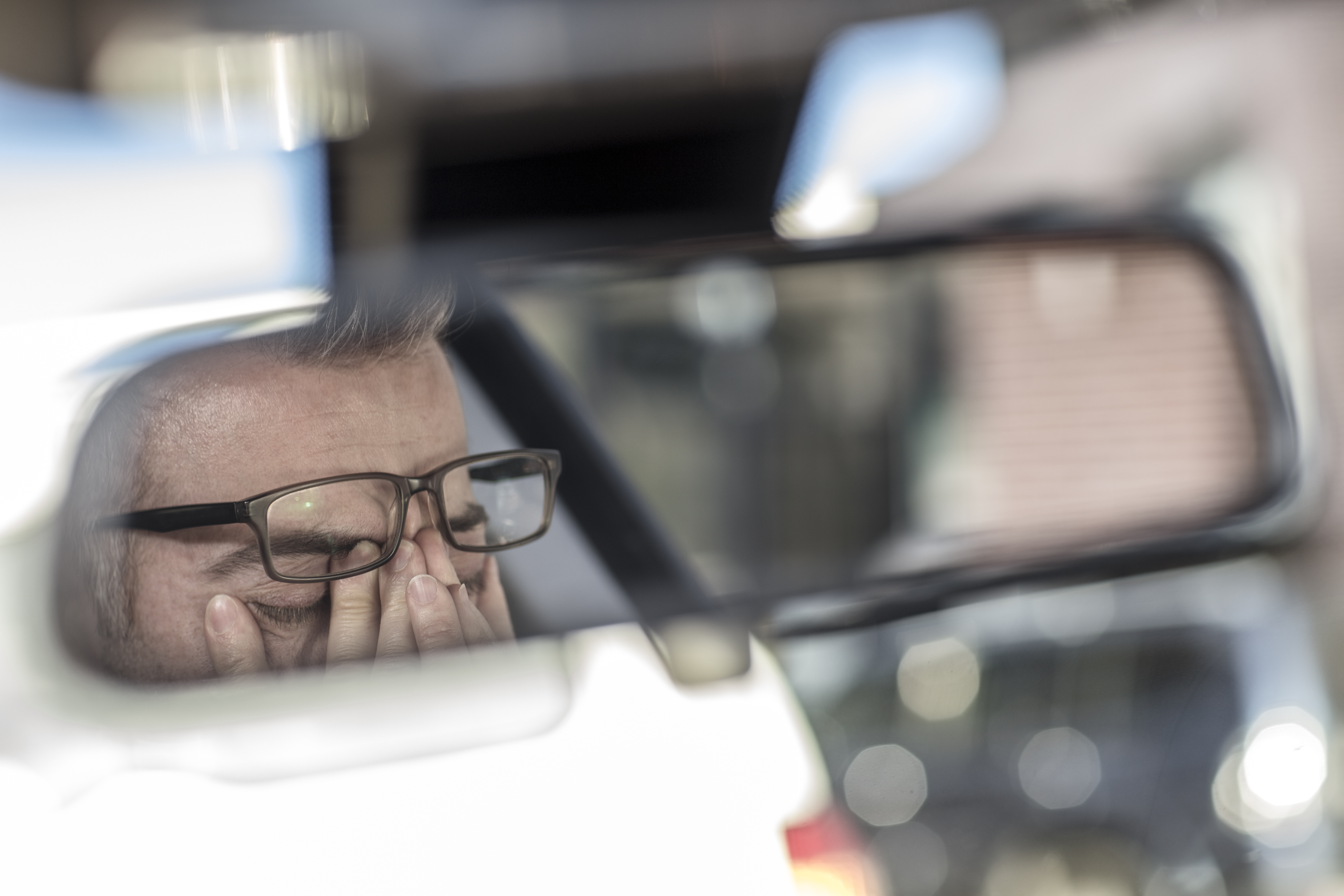man with glasses rubbing his eyes in the rear view mirror of a vehicle